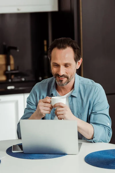 Enfoque selectivo de sonreír hombre maduro utilizando el ordenador portátil y beber café en la cocina - foto de stock
