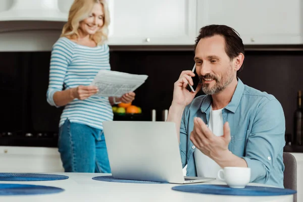 Выборочный фокус улыбающегося мужчины, разговаривающего на смартфоне рядом с ноутбуком, в то время как жена читает газету на кухне — стоковое фото