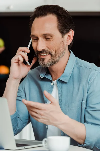 Enfoque selectivo del hombre sonriente hablando en el teléfono celular cerca de la computadora portátil y café en la mesa de la cocina - foto de stock