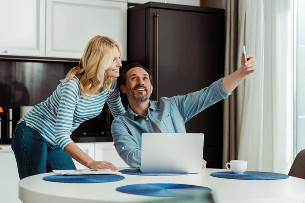 Selective focus of smiling mature couple taking selfie with smartphone in kitchen — Stock Photo