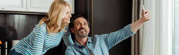 Imagen horizontal de pareja madura sonriente tomando selfie con smartphone en la cocina - foto de stock