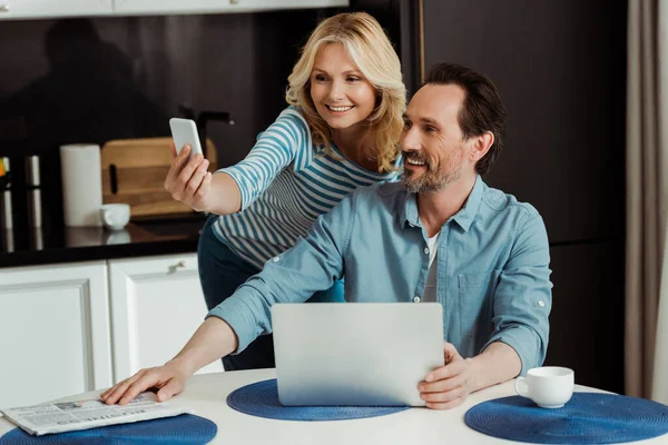 Smiling woman taking selfie with smartphone near husband using laptop in kitchen — Stock Photo