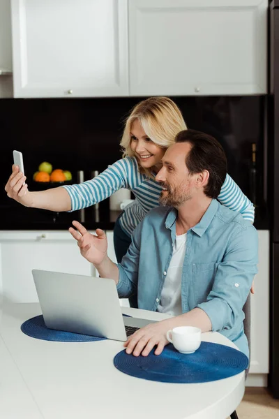 Улыбающаяся женщина делает селфи на смартфоне с мужем возле ноутбука и кофе на кухонном столе — стоковое фото