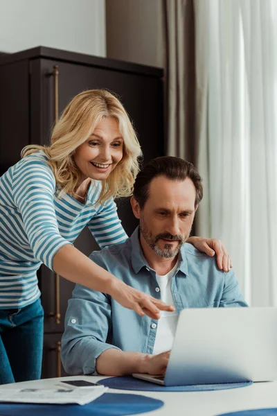 Selective focus of smiling woman pointing with finger at laptop near husband in kitchen — Stock Photo