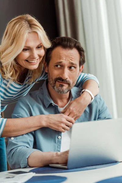 Enfoque selectivo de la mujer sonriente abrazando marido confundido usando ordenador portátil en la cocina - foto de stock