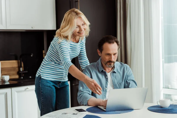 Smiling woman pointing on laptop near husband at kitchen table — Stock Photo