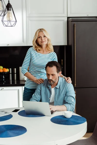 Mature woman looking at camera while hugging husband using laptop in kitchen — Stock Photo
