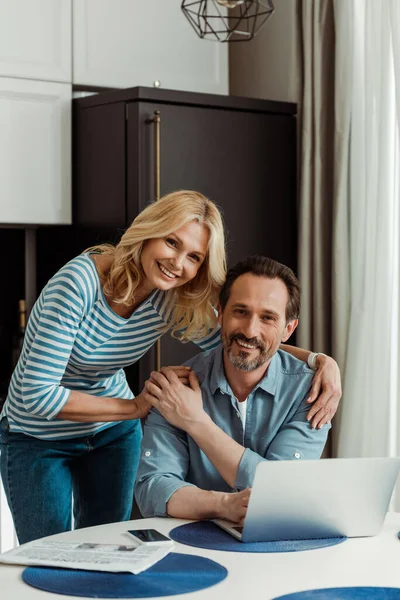 Selective focus of mature woman smiling at camera while embracing husband near digital devices on kitchen table — Stock Photo