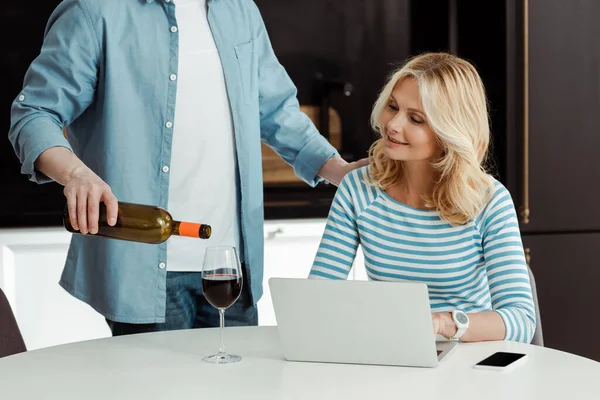 Homem derramando vinho em vidro perto da esposa sorridente usando laptop na mesa da cozinha — Fotografia de Stock