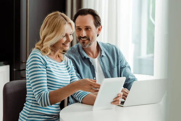 Enfoque selectivo del hombre sonriente usando el ordenador portátil y mirando a la esposa con la tableta digital en la cocina - foto de stock