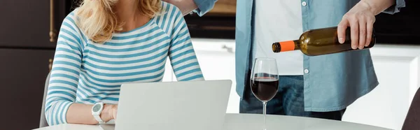 Tiro panorâmico de homem derramando vinho perto da mulher usando laptop na cozinha — Fotografia de Stock