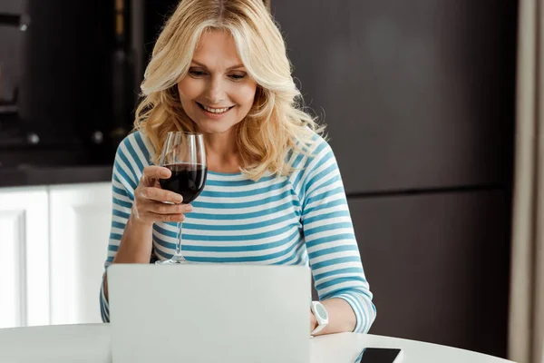 Enfoque selectivo de la mujer sonriente sosteniendo un vaso de vino mientras usa el portátil en la mesa de la cocina - foto de stock