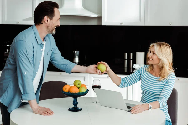 Homme souriant donnant pomme à femme près du vin et ordinateur portable sur la table de cuisine — Photo de stock