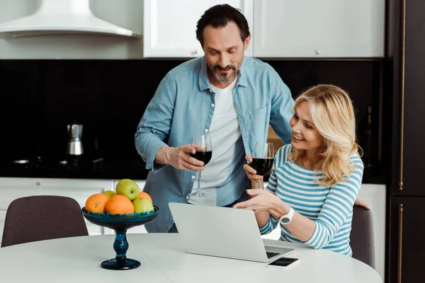 Mujer sonriente sosteniendo un vaso de vino y apuntando a la computadora portátil cerca del marido en la cocina - foto de stock