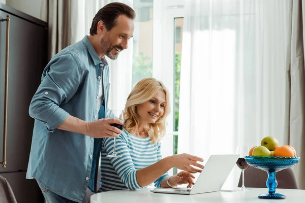 Smiling man holding glass of wine near wife pointing on laptop on kitchen table — Stock Photo