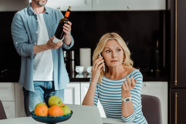 Focus selettivo della donna che parla sullo smartphone vicino al computer portatile mentre l'uomo tiene in mano la bottiglia di vino in cucina — Foto stock