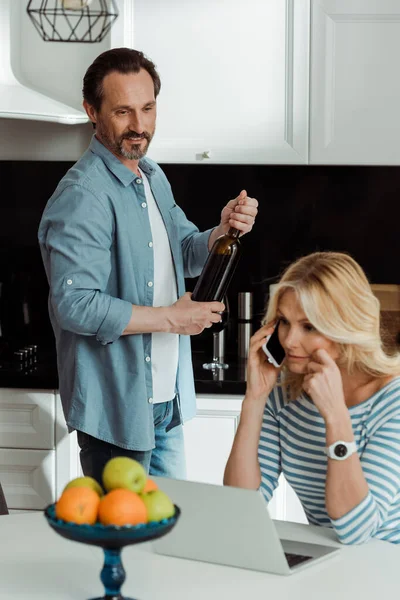 Enfoque selectivo del hombre sonriente sosteniendo la botella de vino cerca de la esposa hablando en el teléfono inteligente en la cocina - foto de stock