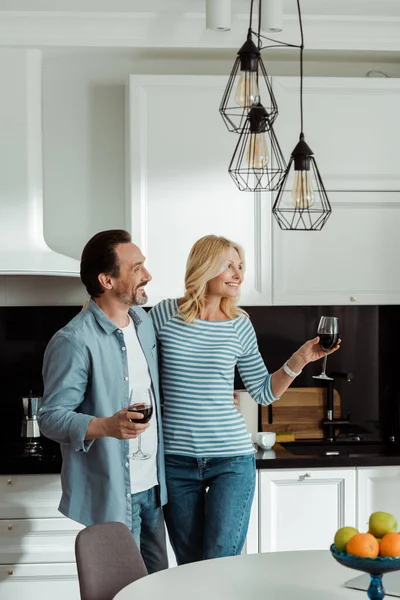 Focus selettivo dell'uomo sorridente che abbraccia la moglie con un bicchiere di vino in cucina — Foto stock