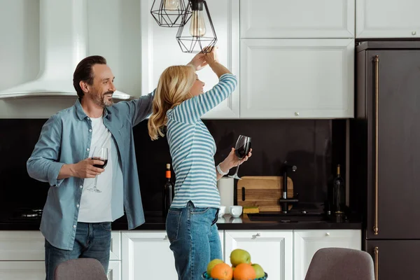 Selective focus of cheerful mature couple dancing with wine glasses in kitchen — Stock Photo