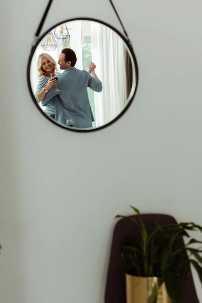 Selective focus of smiling woman reflecting in mirror while holding glass of wine and dancing with husband at home — Stock Photo