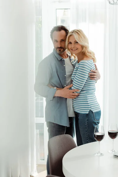 Selective focus of smiling mature couple looking at camera while embracing near glasses of wine in kitchen — Stock Photo