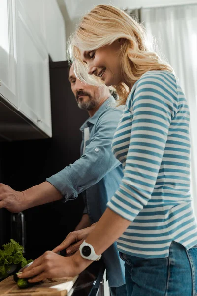 Vista laterale della donna sorridente che taglia il cetriolo vicino al marito in cucina — Foto stock