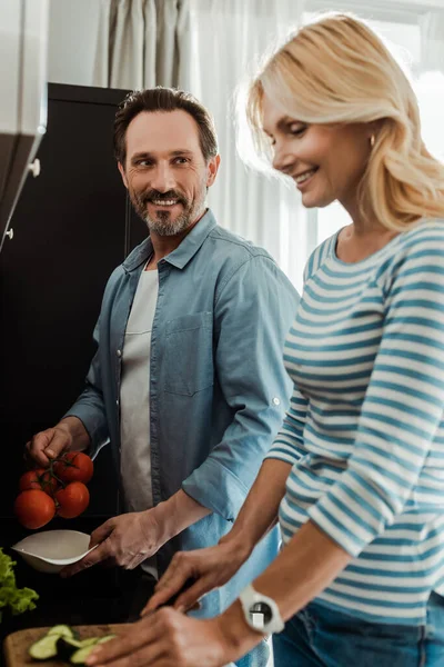 Выборочный фокус улыбающегося мужчины, смотрящего на жену во время приготовления салата на кухне — стоковое фото