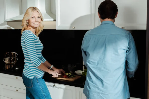 Зріла жінка посміхається на камеру під час різання овочів біля чоловіка на кухні — Stock Photo