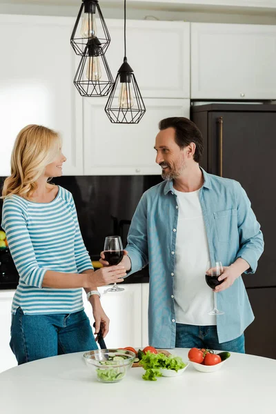 Schöner Mann schenkt lächelnder Frau neben Gemüse auf Küchentisch ein Glas Wein — Stockfoto