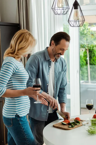 Uomo sorridente che taglia verdure vicino a moglie con vetro di vino in cucina — Foto stock