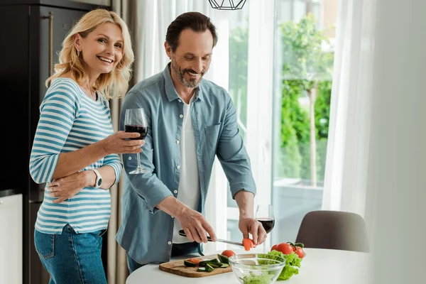 Foco seletivo da mulher com copo de vinho sorrindo para a câmera perto do marido cortando legumes na cozinha — Fotografia de Stock