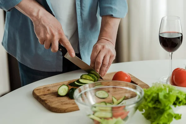 Cropped view of man cutting cucumber while cooking salad near glass of wine in kitchen — Stock Photo