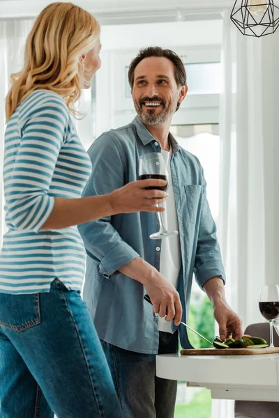 Focus selettivo dell'uomo che sorride alla moglie con un bicchiere di vino durante la cottura dell'insalata in cucina — Foto stock