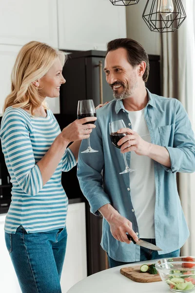 Ältere Paare lächeln einander an, während sie mit Wein in der Nähe von frischem Gemüse auf dem Küchentisch anstoßen — Stockfoto