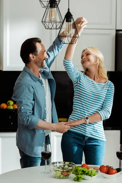 Selective focus of smiling mature couple dancing near salad and wine glasses on kitchen table — Stock Photo