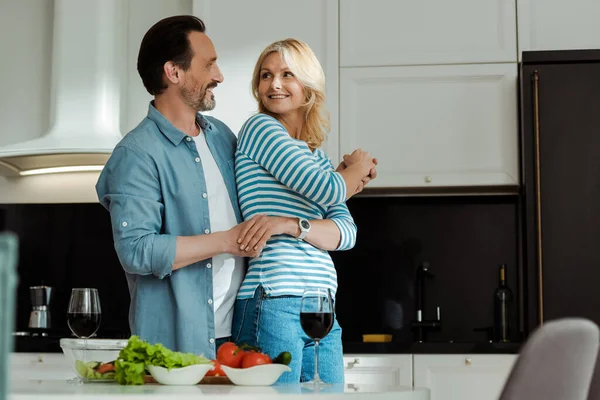 Вибірковий фокус чоловіка обіймає усміхнену дружину біля салату та келихів вина на кухні — стокове фото