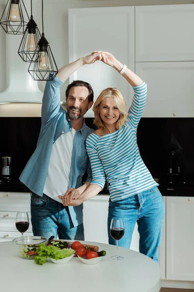 Sonriente pareja madura bailando cerca de ensalada y copas de vino en la mesa de la cocina - foto de stock