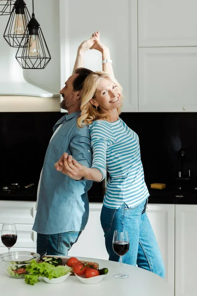 Vista lateral de casal maduro sorridente dançando perto de salada fresca e vinho na mesa da cozinha — Fotografia de Stock