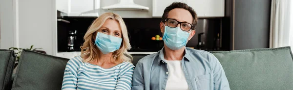 Panoramic shot of mature couple in medical masks sitting on couch at home — Stock Photo
