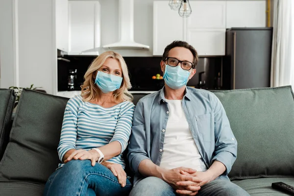 Mature couple in medical masks sitting on couch at home — Stock Photo