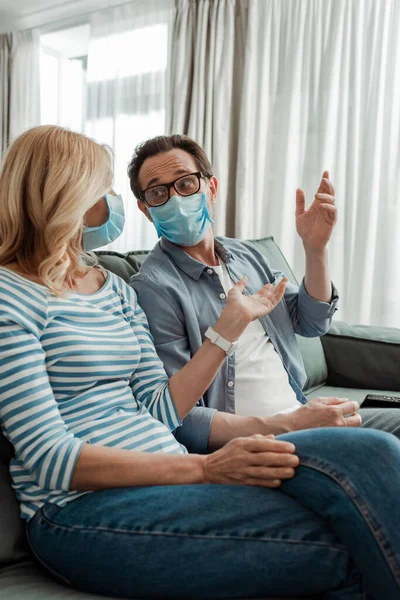 Selective focus of woman in medical mask pointing with hand near husband on couch — Stock Photo