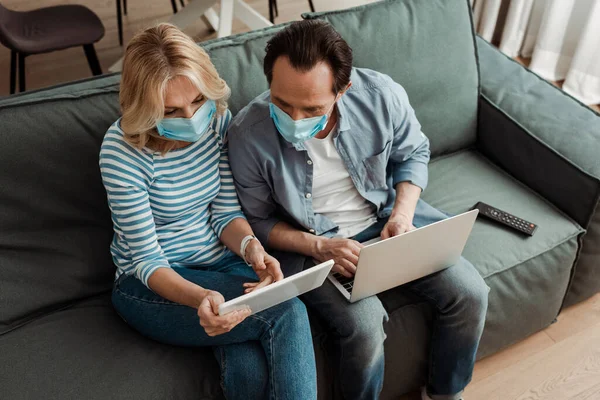 Vista aérea de pareja adulta en máscaras médicas utilizando dispositivos digitales en el sofá - foto de stock