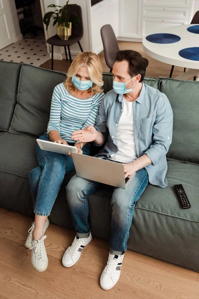 Vista de ángulo alto del hombre en la máscara médica usando el ordenador portátil y apuntando con la mano cerca de la esposa usando la tableta digital en el sofá - foto de stock