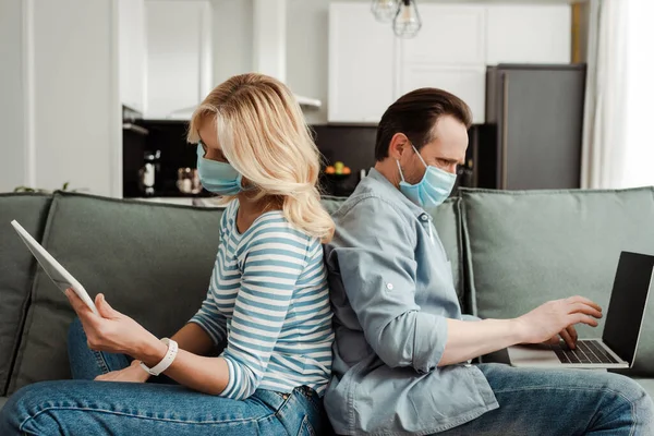 Seitenansicht eines reifen Paares in medizinischen Masken mit Laptop und digitalem Tablet auf der Couch — Stockfoto