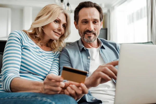 Enfoque selectivo del hombre sonriente apuntando a la computadora portátil cerca de la esposa con tarjeta de crédito en la sala de estar - foto de stock