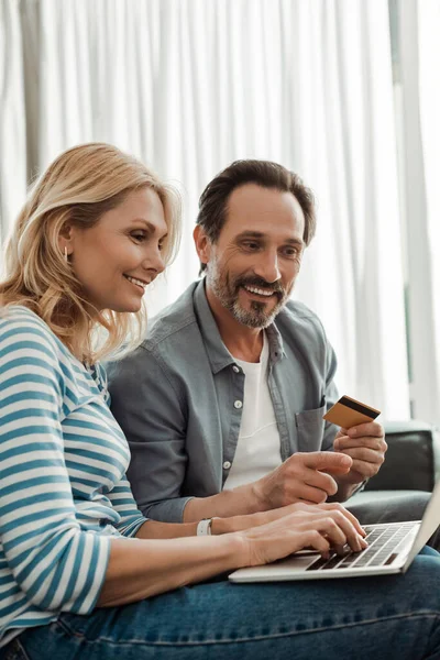 Mujer sonriente usando portátil cerca de marido sosteniendo tarjeta de crédito y señalando con el dedo en casa — Stock Photo