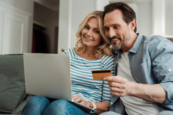 Enfoque selectivo de pareja madura sonriente usando tarjeta de crédito y computadora portátil en el sofá - foto de stock