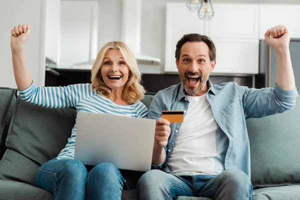 Возбужденная зрелая пара показывает да жест во время использования кредитной карты и ноутбука на диване — стоковое фото