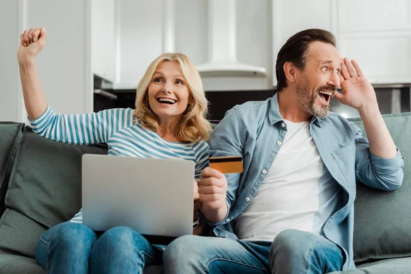 Emocionado casal maduro com laptop e cartão de crédito no sofá em casa — Fotografia de Stock