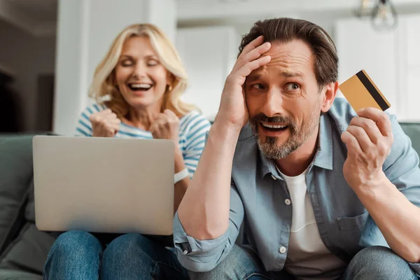 Confundido hombre sosteniendo la tarjeta de crédito cerca de esposa alegre usando el ordenador portátil en casa - foto de stock